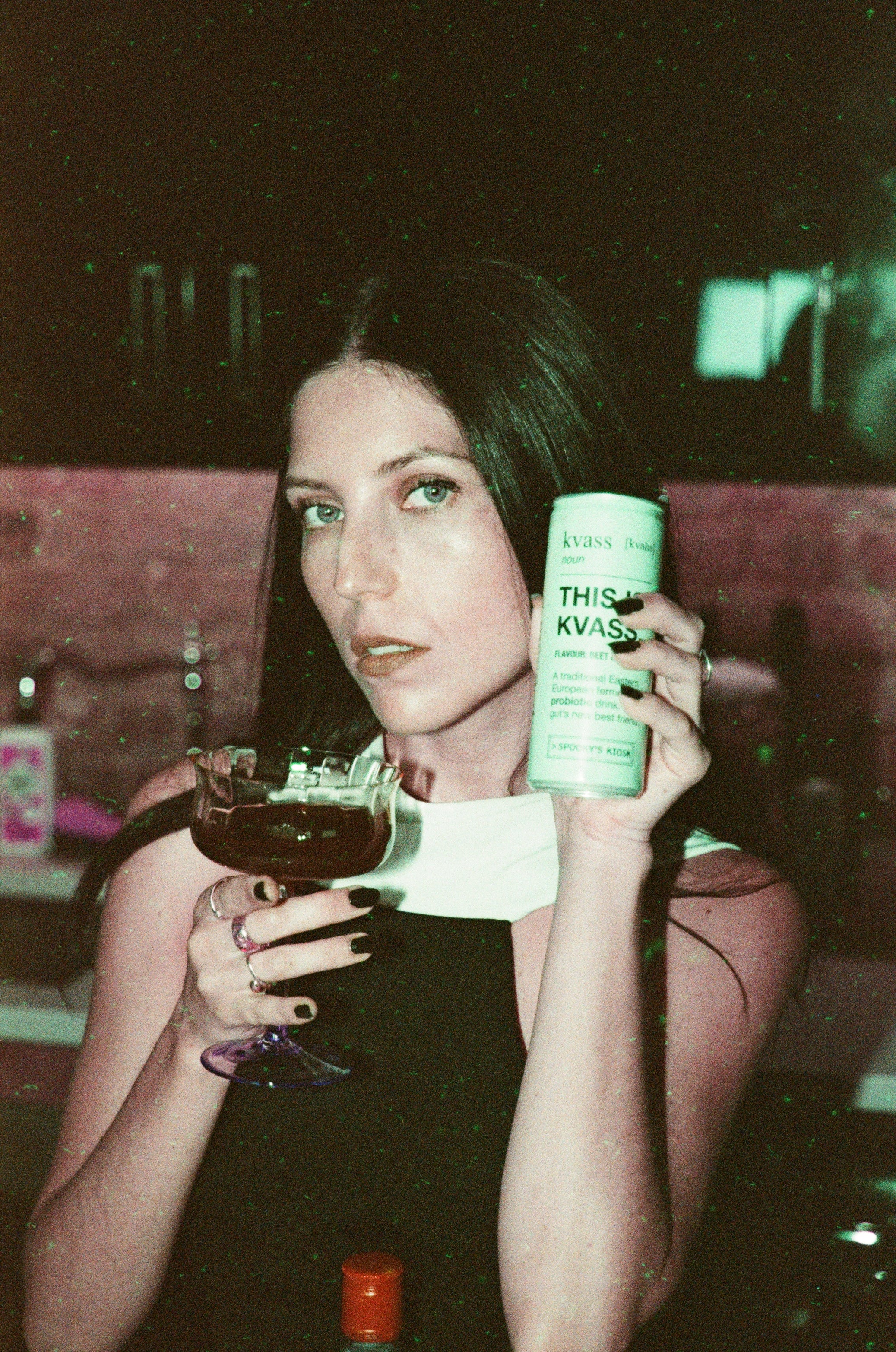 Woman holding a glass of dark liquid and a can of kvass in a dimly lit setting. Avery Wheless oil painting la artist 