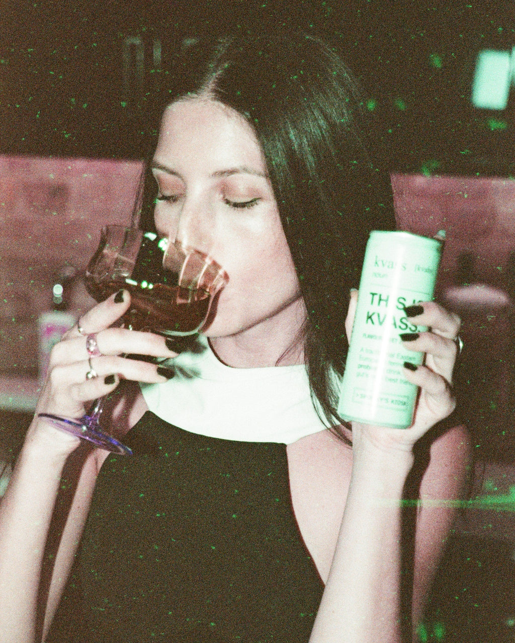 Woman holding a glass of red wine and a can of 'This Is KVASS' in a dark setting