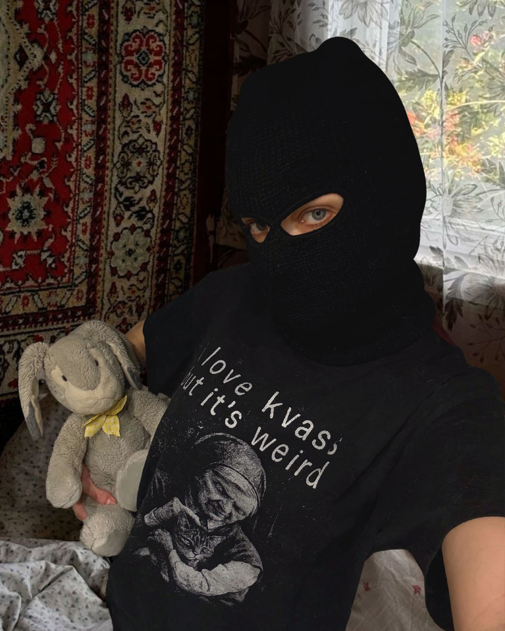 Person wearing a black balaclava and holding a plush toy, with text on a shirt. I love kvass