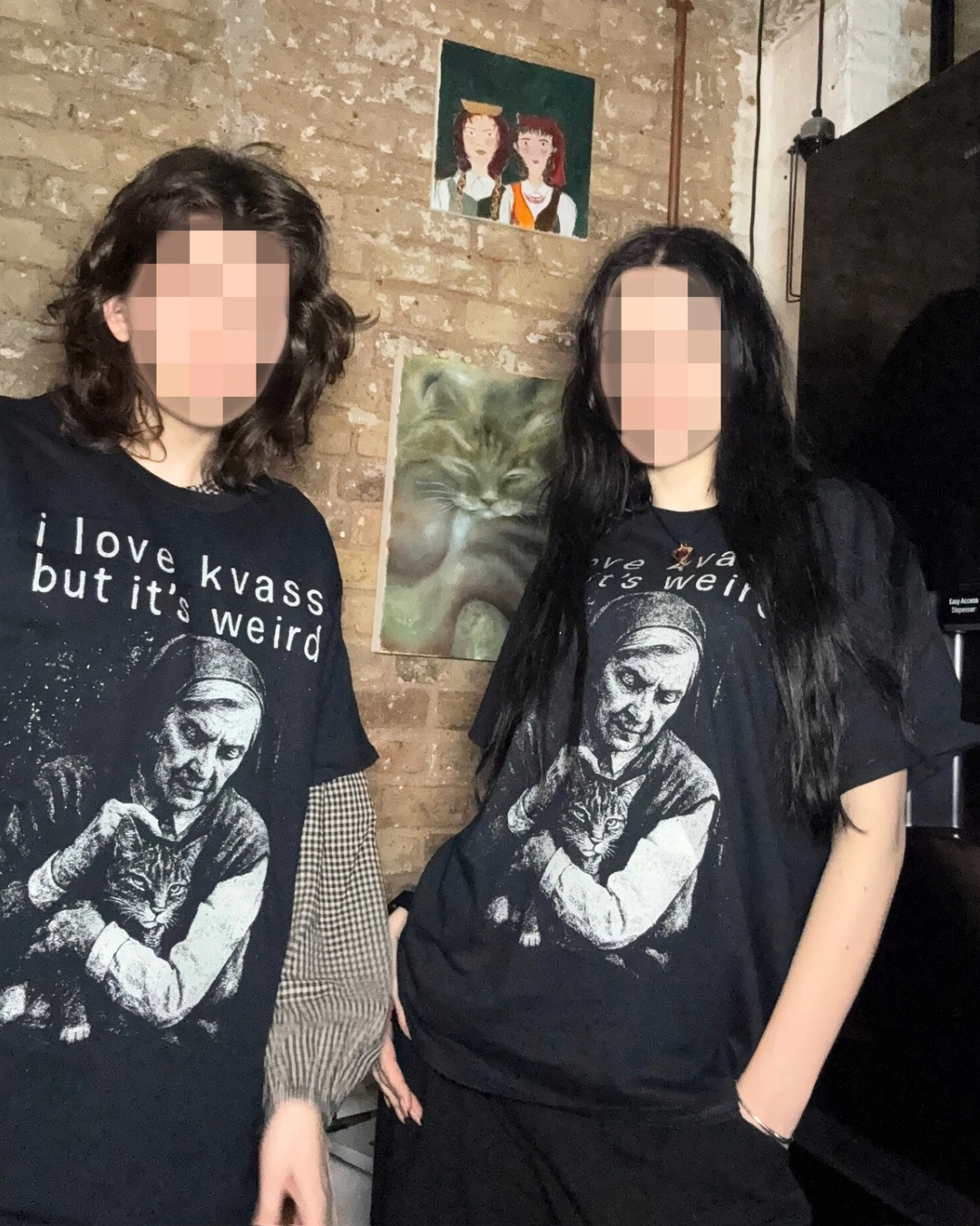 Two people wearing black t-shirts with graphic designs and text, standing against a brick wall with posters.