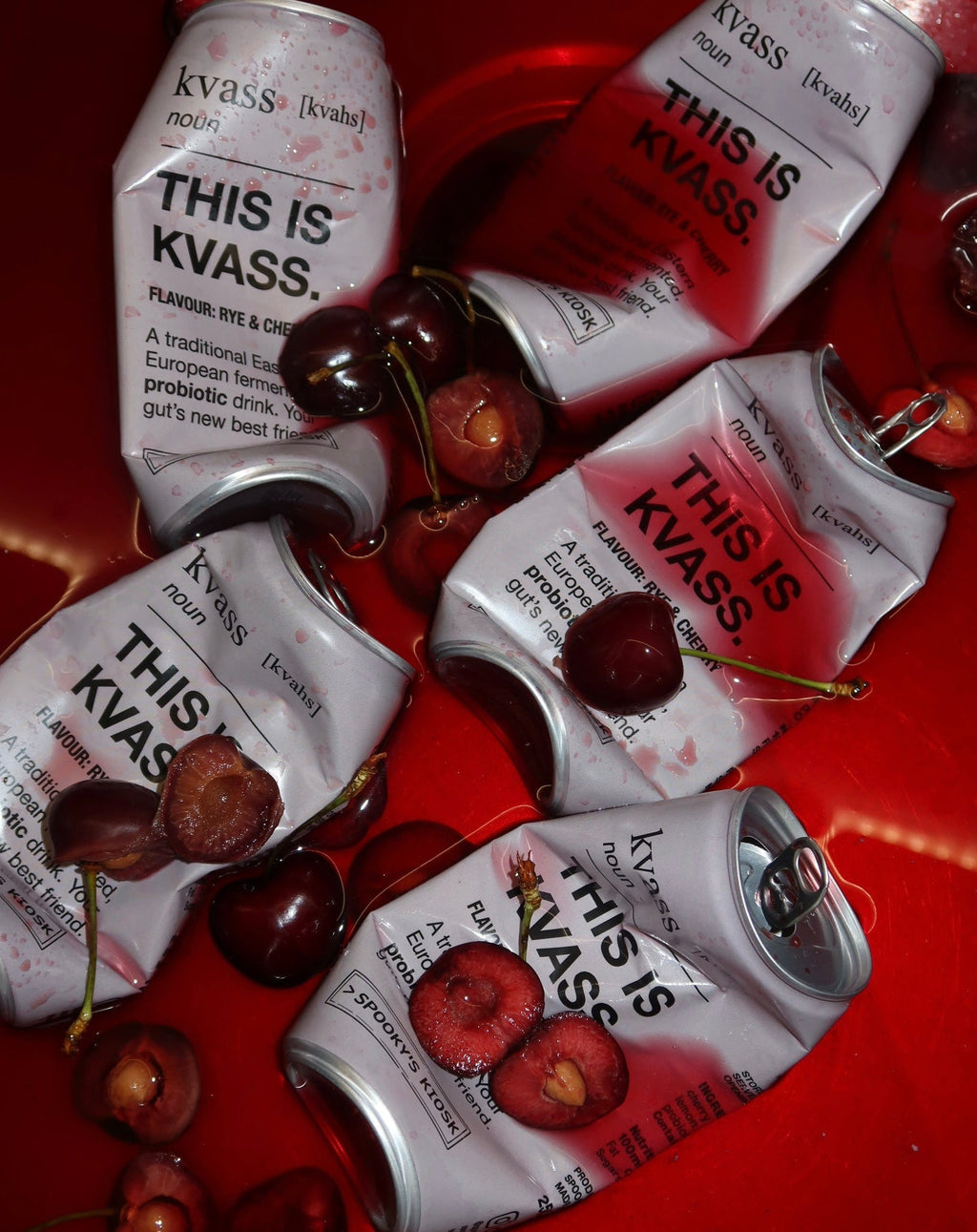 Five silver and red Kvass can drinks are arranged on a glossy red surface with several whole and halved dark cherries scattered around them. The cans show the brand name 'kvass' and the text 'THIS IS KVASS'.