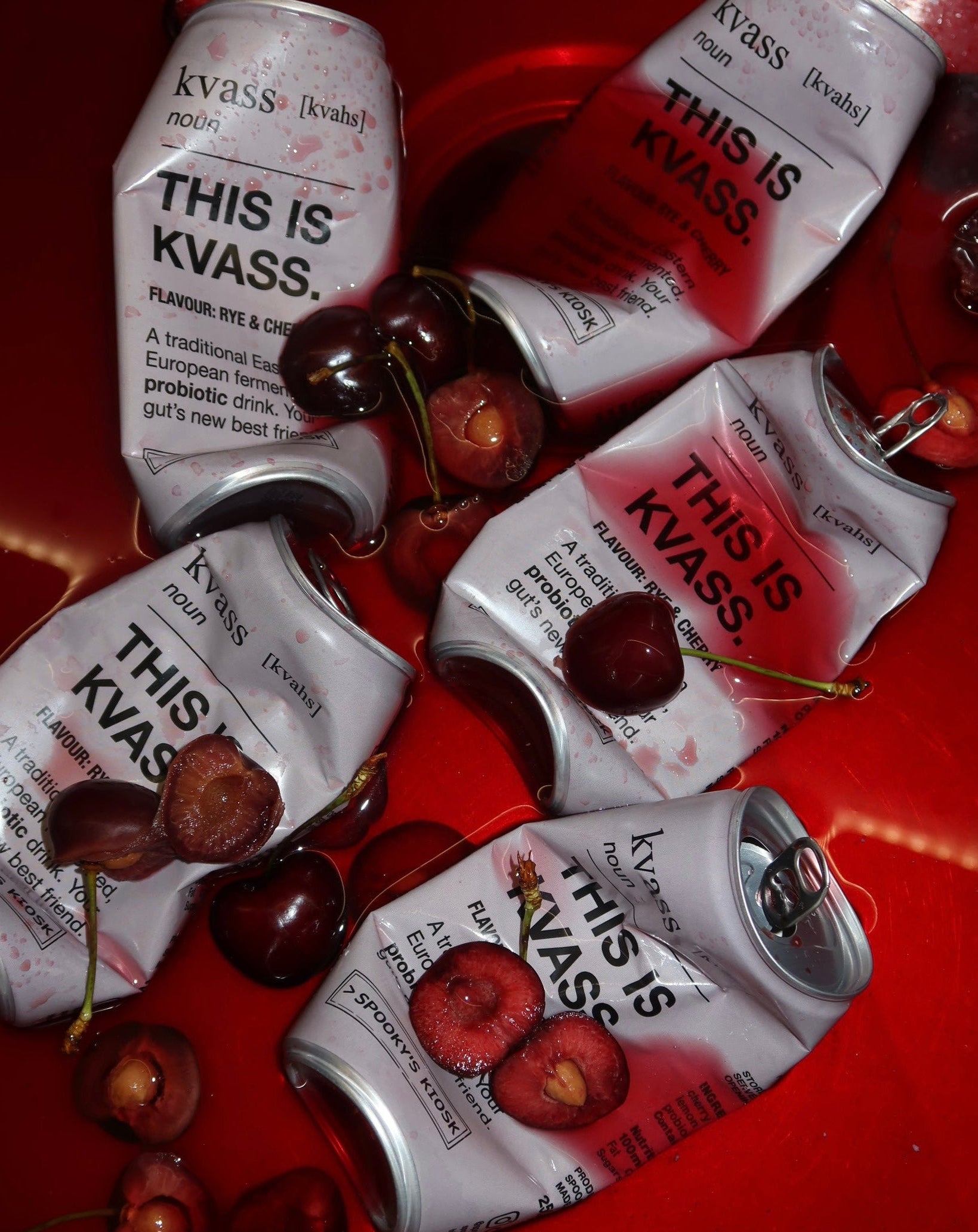 Five silver and red Kvass can drinks are arranged on a glossy red surface with several whole and halved dark cherries scattered around them. The cans show the brand name 'kvass' and the text 'THIS IS KVASS'.