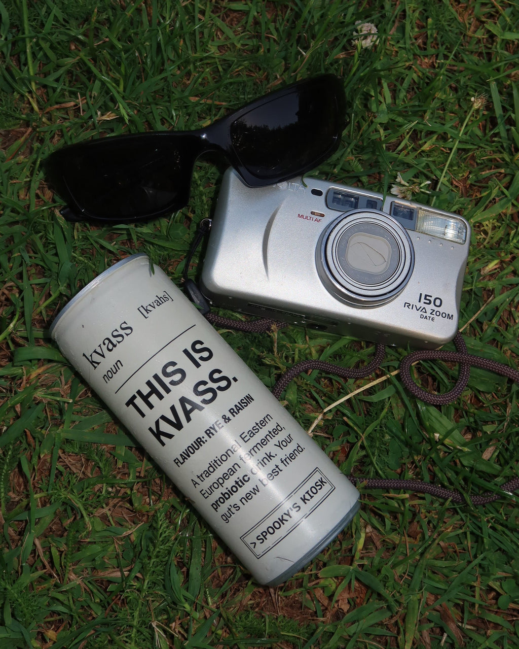 A silver digital camera with a black strap lies on green grass next to black sunglasses and a white cylindrical can printed with the text 'THIS IS KLASSE!'.