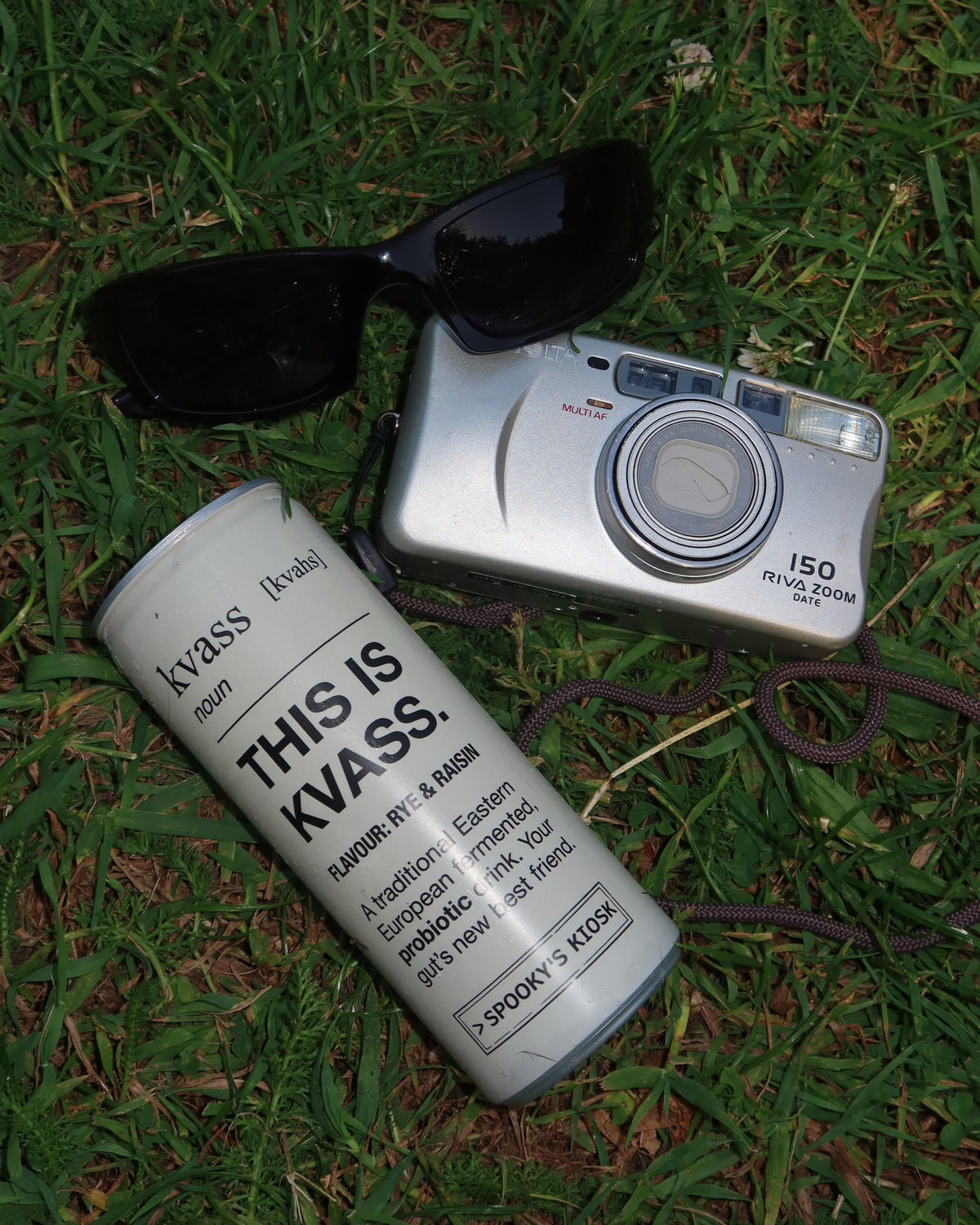 A silver digital camera with a black strap lies on green grass next to black sunglasses and a white cylindrical can printed with the text 'THIS IS KLASSE!'.