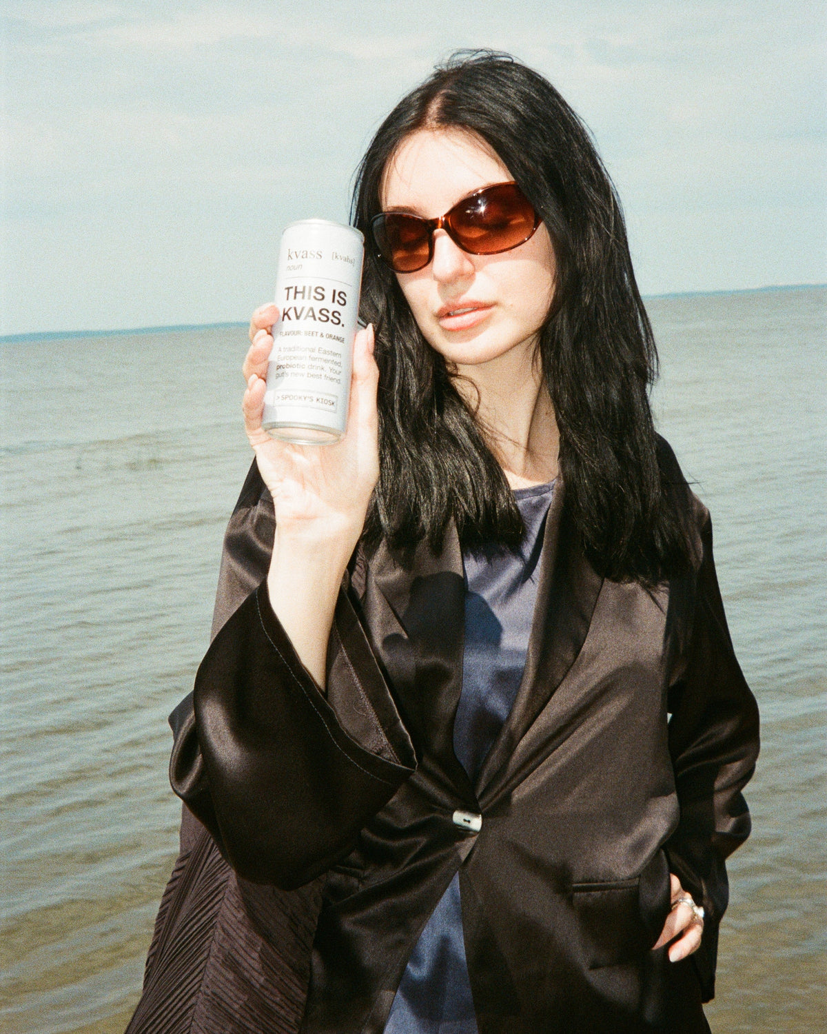 A woman with long black hair and dark sunglasses stands by the water holding a white cylindrical bottle labeled 'THIS IS KVASS'. She is wearing a dark brown satin jacket over a blue top.