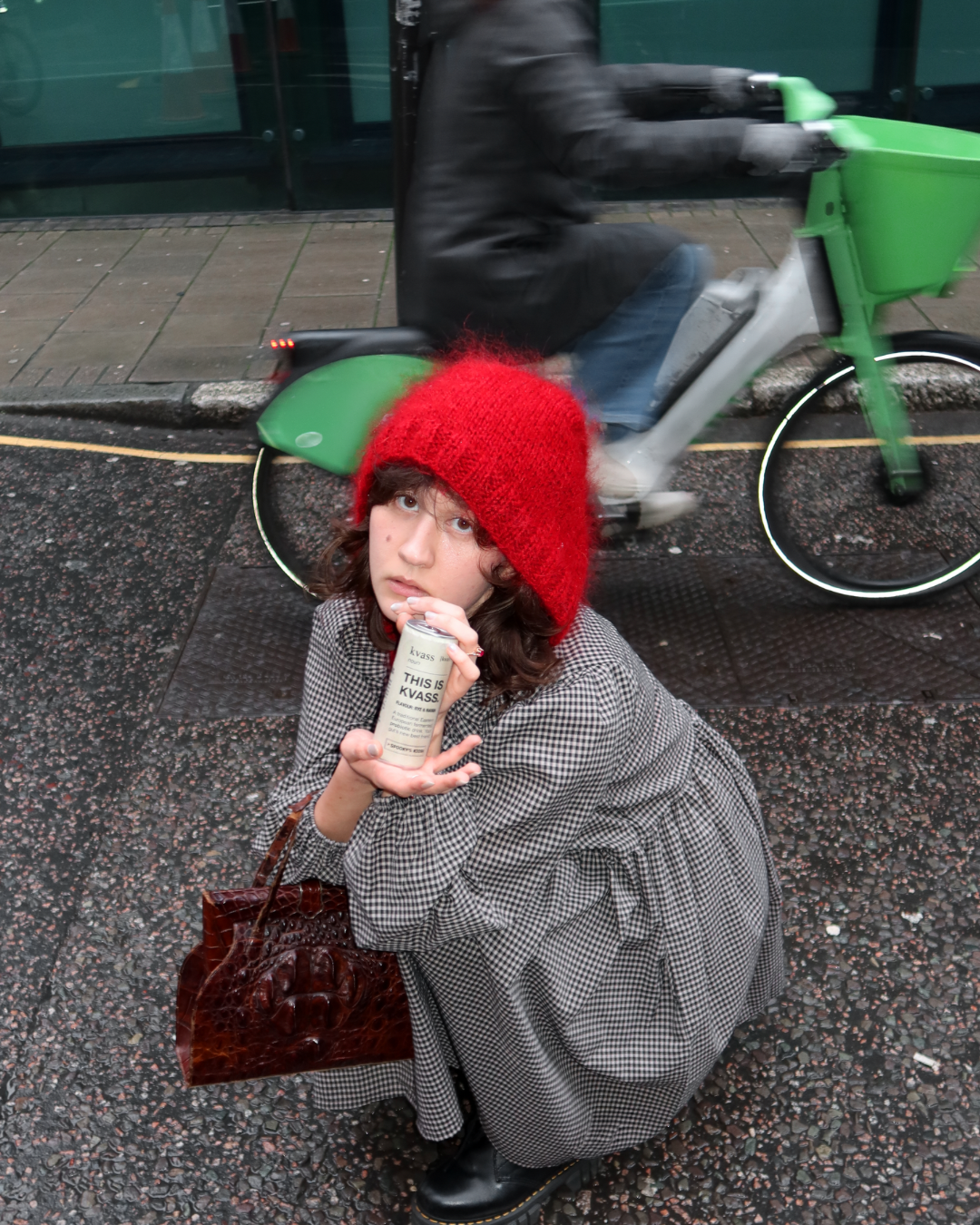 Woman in a red hat and gray coat holding a product on a city street with a cyclist in the background.