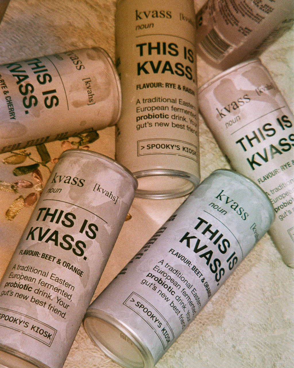 Five glass Kvass cans with printed labels are arranged on a textured beige fabric background. The labels read 'THIS IS KVASS.