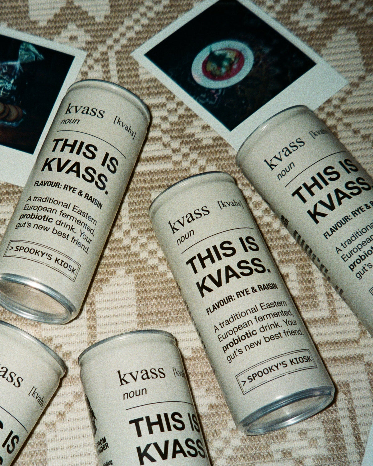Six aluminum cans of kvass arranged on a patterned beige textile with printed polaroid-style photos around them. The cans are light gray with black text reading 'kvass' and 'THIS IS KVASS'.