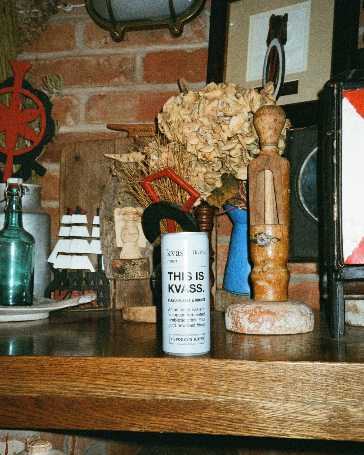 A collection of vintage nautical and maritime-themed decorative items displayed on a wooden shelf, including a white bottle labeled 'THIS IS KVASS', a wooden ship's wheel, a green glass bottle, a small ship model, and framed art against a brick wall background.