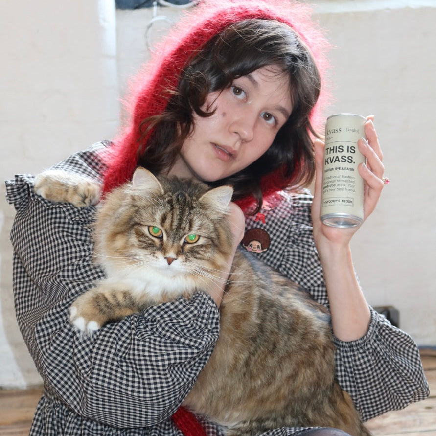 Person holding a cat and a can of kvass, wearing a red hood and plaid coat.
