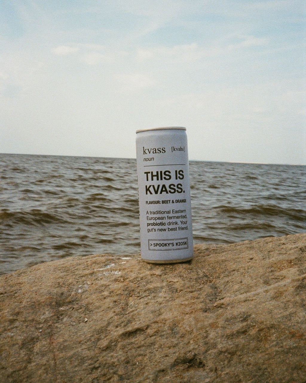 A blue can of Kvass beverage standing upright on a rocky shoreline with water and a pale blue sky in the background.