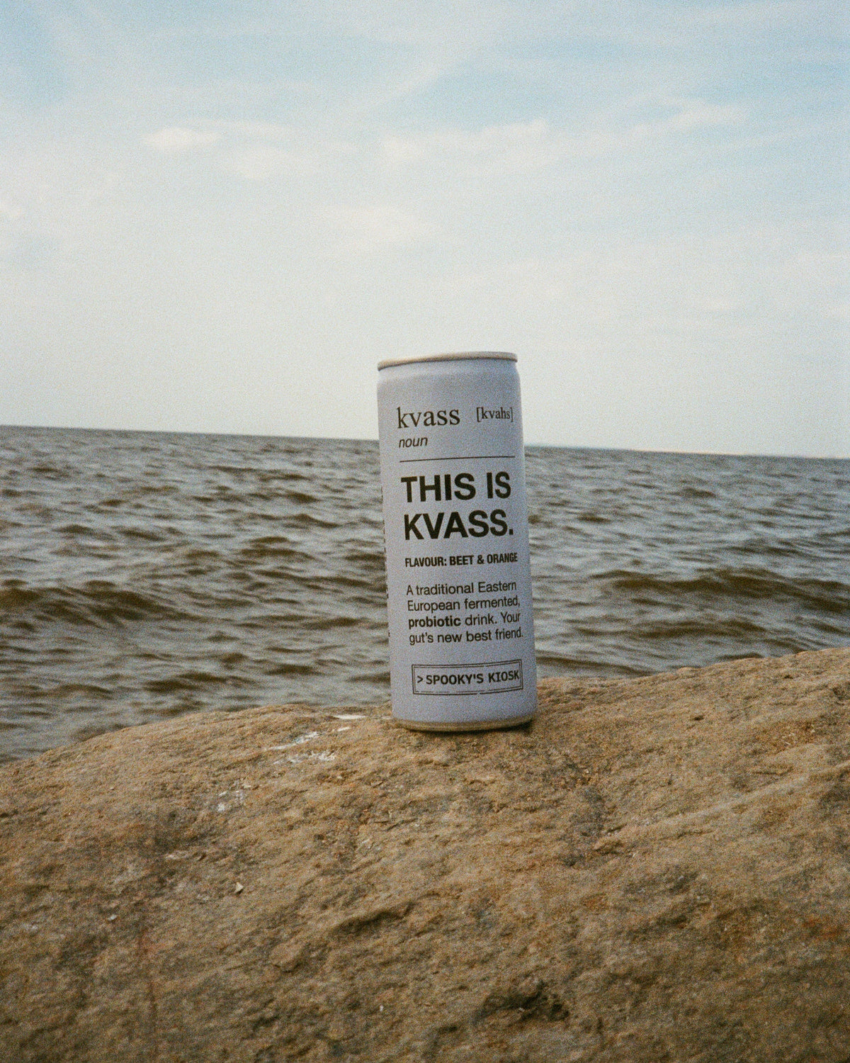 A blue can of Kvass beverage standing upright on a rocky shoreline with water and a pale blue sky in the background.