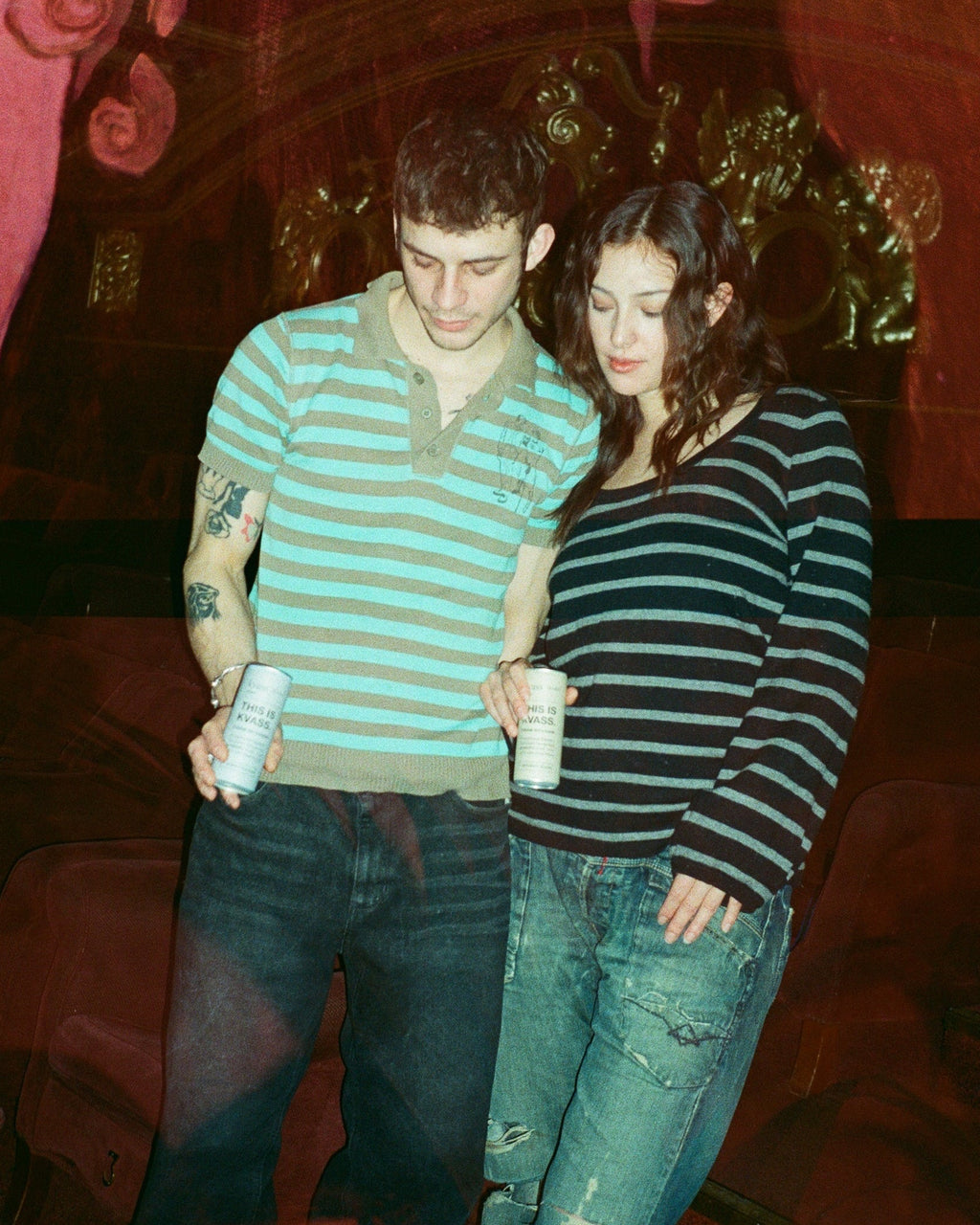 Two people standing together holding kvass cans in a dimly lit setting