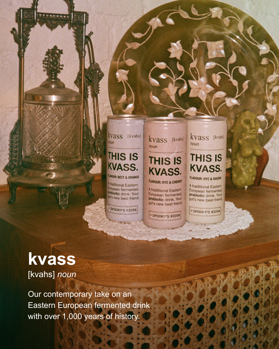 Three cans of kvass beverage arranged on a wooden table with a lace doily, alongside decorative objects including a metal lantern and a carved plate. Text at the bottom reads 'kvass [kvahs] noun'.