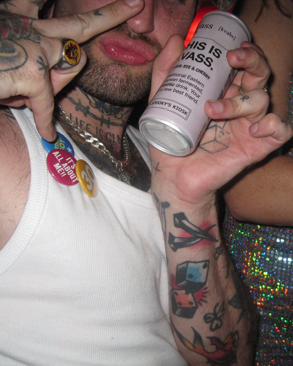 A close-up party scene showing a tattooed man in a white tank top holding a can of 'This Is kvass' can, with another person in a silver rhinestone dress beside him.