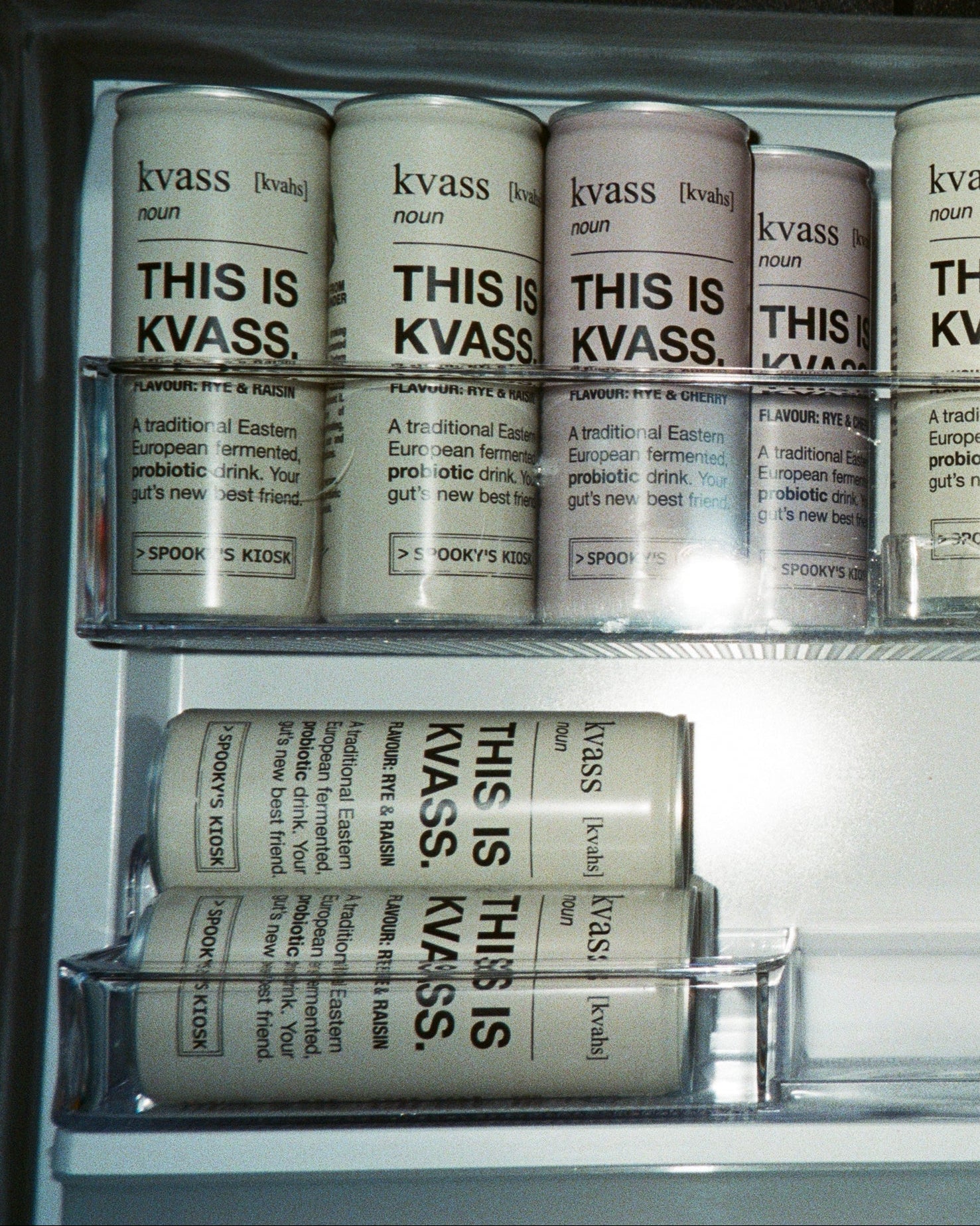 A set of cylindrical beverage cans with white labels and black text reading 'kvass' and 'THIS IS KVASS' arranged inside a clear glass refrigerator or cooler shelf.