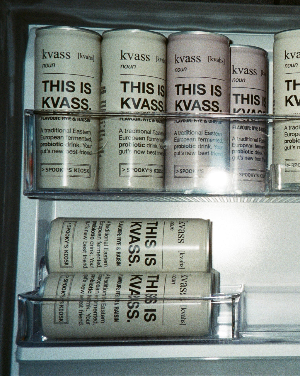 A set of cylindrical beverage cans with white labels and black text reading 'kvass' and 'THIS IS KVASS' arranged inside a clear glass refrigerator or cooler shelf.