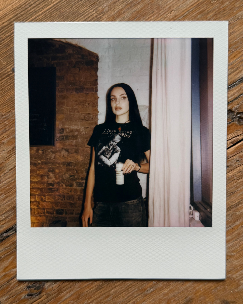 A Polaroid-style instant photo print lying on a wooden surface, showing a person standing indoors between a brick wall and a curtain, wearing a black graphic T-shirt and holding a kvass can